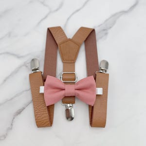 Könnte beinhalten: Ein Set brauner Lederhosenträger mit einer rosafarbenen Fliege. Die Hosenträger haben silberne Clips und die Fliege ist aus weichem Stoff.