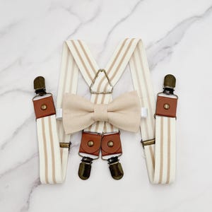 Puede incluir: Un conjunto de tirantes a rayas beige y blancas con detalles en cuero marrón y una pajarita beige a juego.