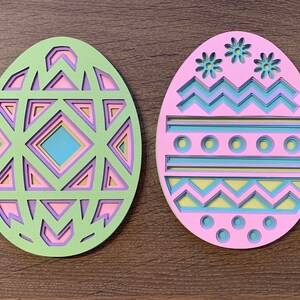 Può includere: Due uova di Pasqua di carta color pastello con intricati disegni ritagliati. Un uovo presenta un motivo geometrico in tonalità di rosa, verde e blu. L'altro uovo ha un motivo a chevron, strisce e cerchi in tonalità di rosa, blu e giallo.