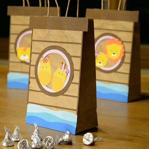 May include: Brown paper gift bags with animal illustrations, including giraffes and lions, set against a blue wave design. Each bag has a brown handle and is decorated with a circular frame. Silver foil-wrapped chocolate kisses are scattered in front.