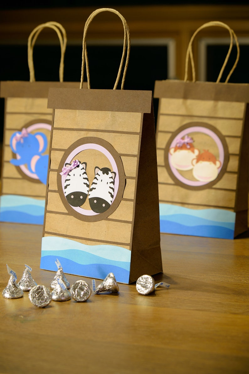 May include: Brown paper gift bags with animal-themed cutouts, including zebras, elephants, and monkeys. Each bag has a blue wave design at the bottom and a brown handle. Silver chocolate candies are scattered in front of the bags.
