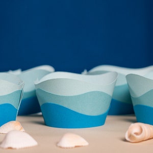 May include: Blue and white paper cupcake liners with a wave design. The liners are arranged in a row on a table with seashells scattered in front of them.