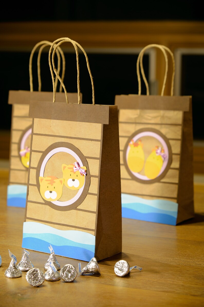 May include: Brown paper gift bags with a decorative design. Each bag features a circular design with cartoon tiger faces and a blue wave pattern at the bottom. Several foil-wrapped chocolate candies are in front of the bags.