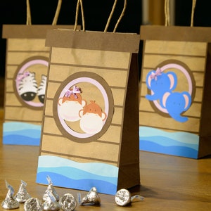 May include: Brown paper gift bags with animal-themed cutouts. Each bag features a different animal: a zebra, two monkeys, and a blue elephant. The bags have blue wave accents and brown handles. Silver foil-wrapped chocolate candies are in front.