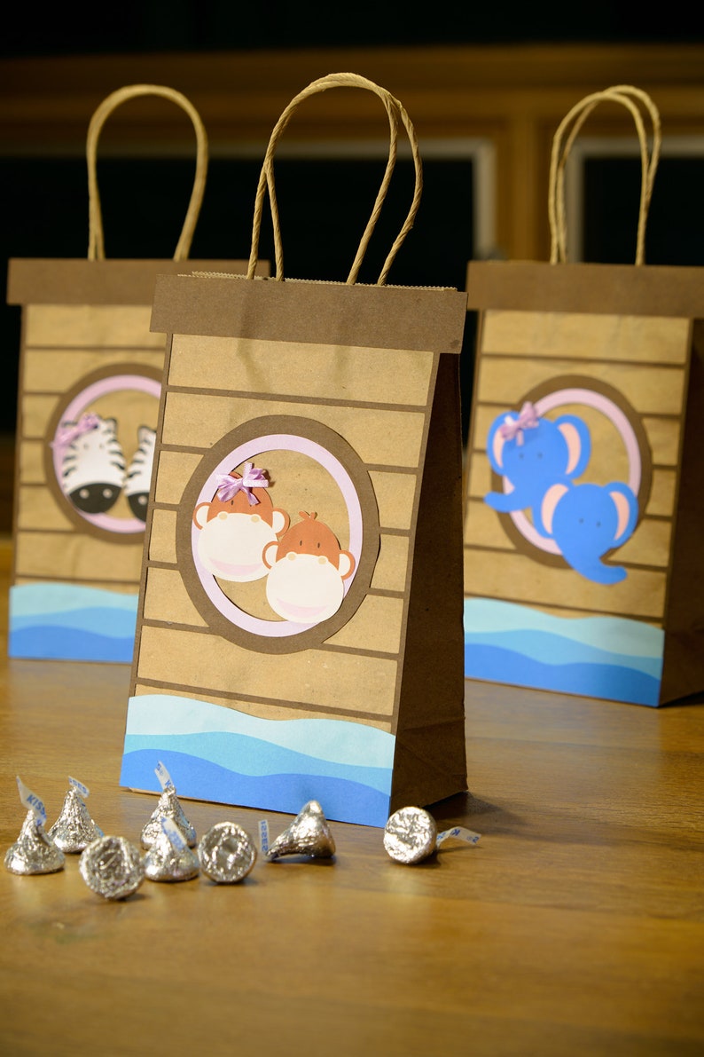 May include: Brown paper gift bags with animal-themed cutouts. Each bag features a different animal: a zebra, two monkeys, and a blue elephant. The bags have blue wave accents and brown handles. Silver foil-wrapped chocolate candies are in front.