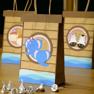May include: Brown paper gift bags with animal-themed cutouts, including elephants and zebras, and blue wave accents. Each bag has a brown handle and is decorated with a circular design. Silver foil-wrapped chocolate candies are in front of the bags.