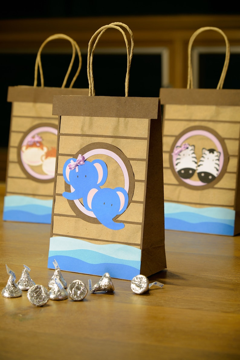 May include: Brown paper gift bags with animal-themed cutouts, including elephants and zebras, and blue wave accents. Each bag has a brown handle and is decorated with a circular design. Silver foil-wrapped chocolate candies are in front of the bags.