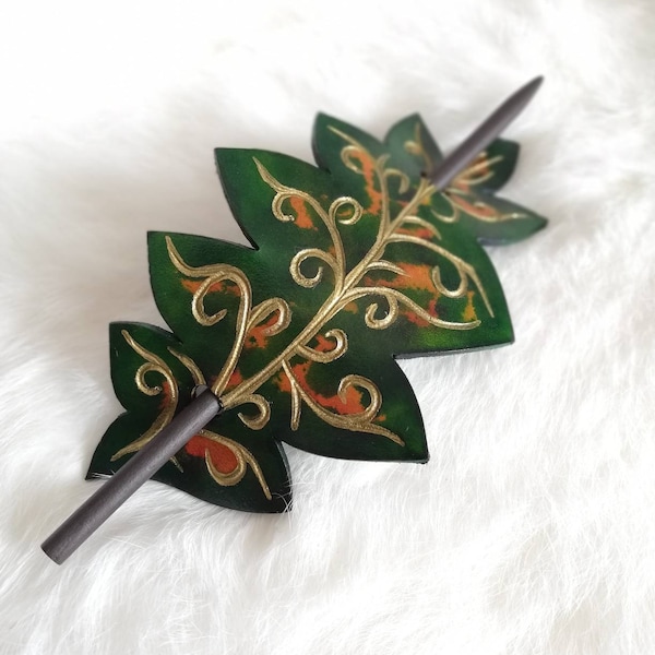 Puede incluir: Horquilla para el cabello artesanal con forma de hoja. La horquilla es verde con detalles dorados y naranjas, y un palito marrón oscuro. El diseño de la hoja presenta intrincados remolinos y patrones dorados. Un accesorio para el cabello único.