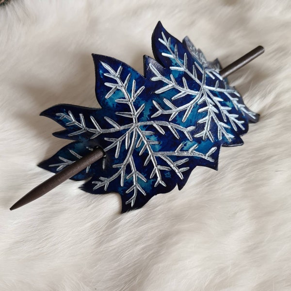 Puede incluir: Una pinza para el pelo de cuero azul con un diseño de helecho plateado y un palo de madera. La pinza está sobre una superficie de pelo blanco.