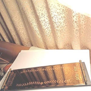 May include: A rectangular serving tray with a mirrored surface and gold-colored floral and leaf design. The tray has gold handles on each end and a dark brown border. The background features a beige curtain with a floral pattern.