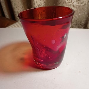 May include: A vibrant red glass tumbler featuring a flared rim and a tapered base. The glass has a smooth, translucent finish with subtle internal air bubbles and a slightly irregular, organic shape throughout the body of the vessel.