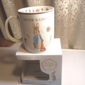 May include: A white ceramic mug featuring a gold-toned rim and an illustration of Peter Rabbit in a blue jacket. The text PETER RABBIT is printed on the front. The mug sits on a matching cardboard box with a cutout and the text PETER RABBIT.