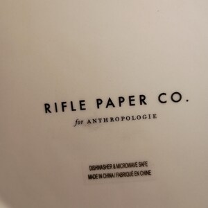 May include: Close-up of a white ceramic surface with the text "RIFLE PAPER CO." in black, followed by "for ANTHROPOLOGIE." Below, a sticker reads "DISHWASHER & MICROWAVE SAFE MADE IN CHINA/FABRIQU&Eacute; EN CHINE."