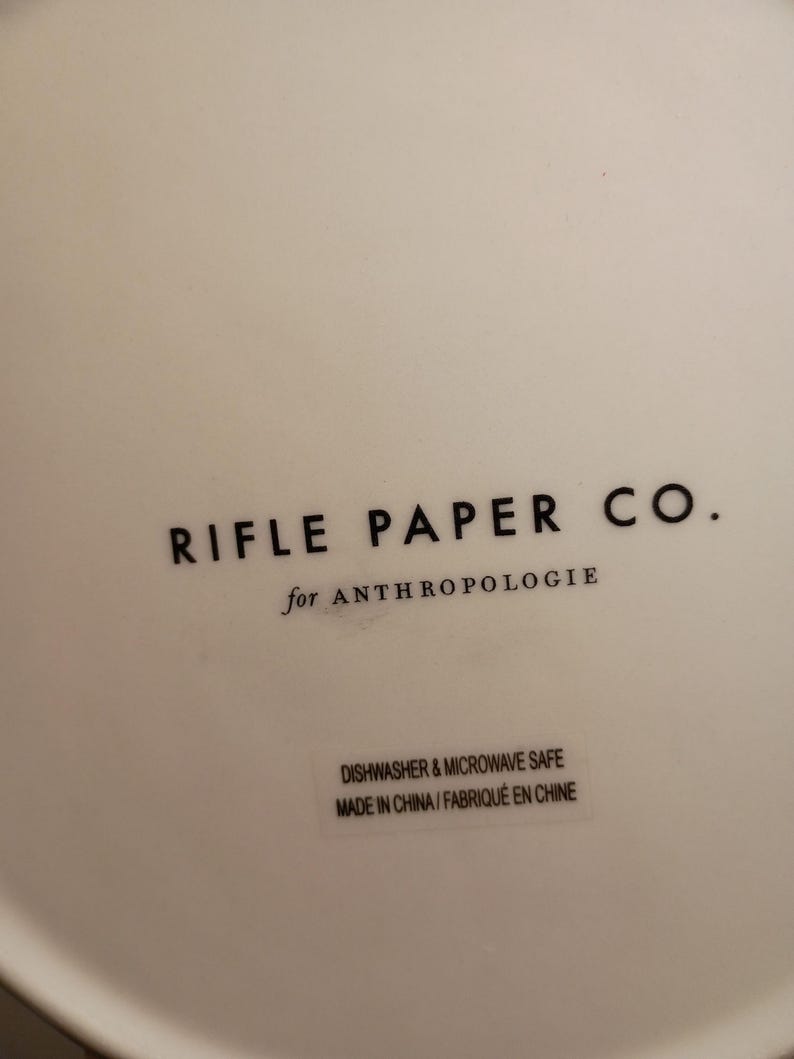 May include: Close-up of a white ceramic surface with the text "RIFLE PAPER CO." in black, followed by "for ANTHROPOLOGIE." Below, a sticker reads "DISHWASHER & MICROWAVE SAFE MADE IN CHINA/FABRIQU&Eacute; EN CHINE."