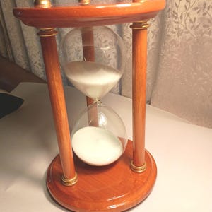 May include: A wooden hourglass with a clear glass center filled with white sand. The hourglass has a round base and top, connected by four wooden pillars. Gold-colored accents are visible on the top and bottom.