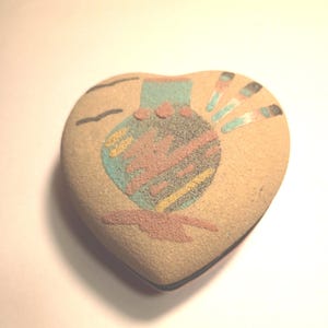 May include: Heart-shaped decorative stone with a Native American design. The design features a turquoise vase with red, yellow, and black accents. The stone is tan with a smooth texture.