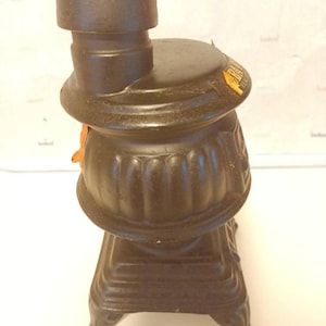May include: A brown ceramic bottle shaped like a wood-burning stove with a black metal chimney. The bottle has a label that reads "The Old Stove."