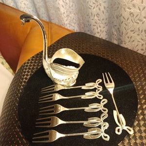 May include: A set of six silver-toned dessert forks with decorative bows on the handles, arranged on a black velvet surface. A silver swan-shaped dish is in the background.