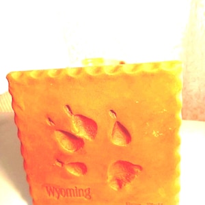 May include: A square, orange-toned plaque with scalloped edges featuring an embossed animal paw print in the center. The word Wyoming is printed in the lower left corner, and the text Gray Wolf is visible in the lower right corner.