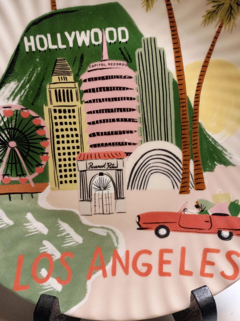 May include: A decorative plate featuring a colorful illustration of Los Angeles landmarks. The plate includes the Hollywood sign, Capitol Records, a Ferris wheel, and a red convertible car. The words "HOLLYWOOD" and "LOS ANGELES" are prominently displayed.