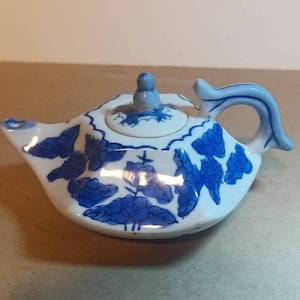 May include: A small white teapot with a blue and white floral design. The teapot has a lid and a handle.