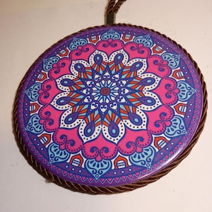 May include: A circular wall hanging featuring a vibrant mandala pattern in shades of pink, purple, light blue, and orange. The disc is edged with a thick, twisted brown rope trim and includes a matching brown rope loop at the top for hanging.