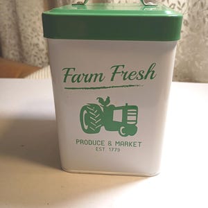May include: A white metal container with a green lid and handle. The words "Farm Fresh" are printed on the front, along with a green tractor graphic and the text "Produce & Market EST. 1779".