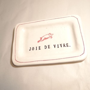 May include: A rectangular white ceramic tray with rounded corners, featuring a red dotted border, a small red illustration of a jumping pig, and the black text "JOIE DE VIVRE." printed in the center of the tray surface.