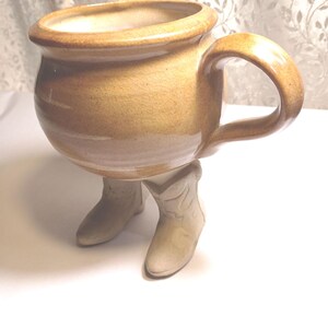 May include: A ceramic mug with a glossy tan glaze and a rounded body, featuring a single handle on the side. The base of the mug is uniquely designed to resemble two light-colored cowboy boots that serve as the feet for the vessel.