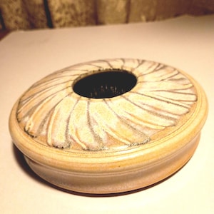 May include: A ceramic container with a circular shape and a central hole. The lid features a pattern of radiating lines in shades of beige and gray. The container's base is a solid beige color, creating a contrast with the lid's design.