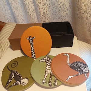 May include: Set of five round coasters featuring animal illustrations. Includes a giraffe, zebra, ostrich, and monkey. The coasters are in shades of orange, green, and brown, with a black box for storage.