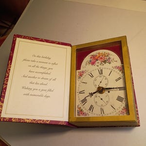 May include: A decorative clock in a book-shaped frame. The clock face is white with black Roman numerals and floral accents. The open book displays a birthday message on the left page. The frame is gold-colored.