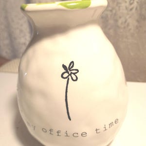 May include: White ceramic vase with a rounded body and a flared top. The rim is accented with green paint. A black floral design and the words "my office time" are printed on the front. A decorative item for home or office.