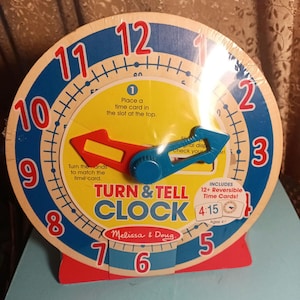 May include: A wooden clock with colorful numbers and hands. The clock has a blue and red border and a yellow face. The clock is labeled "Turn & Tell Clock" and "Melissa & Doug". The clock comes with 12 reversible time cards. The packaging includes the text "Includes 12+ Reversible Time Cards! 4:15 Ages 4+ "