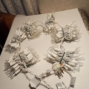 May include: A collection of white Christmas lights, bundled together with green ties. The lights have clear bulbs and white wiring. A power plug and a white connector are visible, along with caution labels.