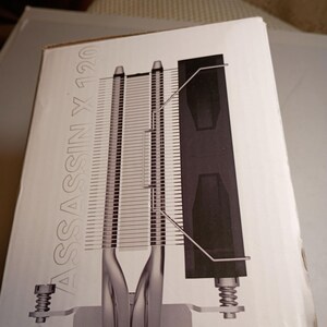 May include: A white box with a diagram of a computer CPU cooler. The cooler has silver fins, a black heat sink, and the text "ASSASSIN X 120" in silver. The cooler is designed to dissipate heat from a computer's central processing unit.