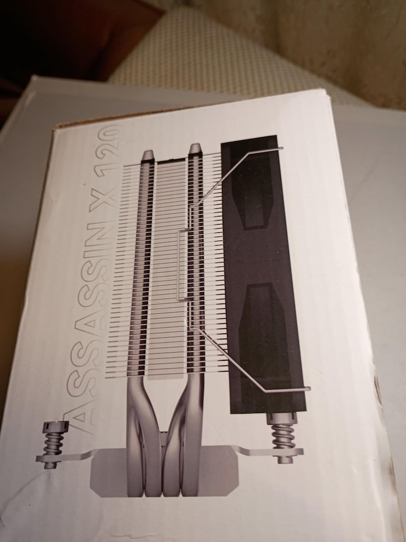 May include: A white box with a diagram of a computer CPU cooler. The cooler has silver fins, a black heat sink, and the text "ASSASSIN X 120" in silver. The cooler is designed to dissipate heat from a computer's central processing unit.