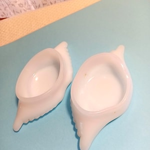 May include: Two white, seashell-shaped dishes with a smooth, glossy finish. The dishes have a rounded bowl and a pointed, textured base. They are set against a light blue background.