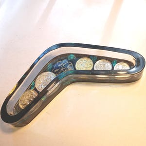 May include: A boomerang-shaped resin tray with a clear, iridescent finish. The tray contains several coins and decorative elements. The coins are silver and gold-colored. The tray is on a white surface.
