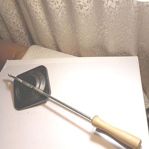 May include: A vintage black metal sandwich toaster with a long, silver handle and a light-colored wooden grip. The toaster has a square cooking surface and a hinged design for easy use. A classic kitchen tool.