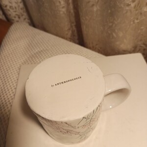 Puede incluir: Taza de cer&aacute;mica blanca con un dise&ntilde;o floral en rosa y verde. La taza tiene un asa curva y el texto "by ANTHROPOLOGIE" impreso en la parte inferior. La taza est&aacute; sobre una superficie blanca.