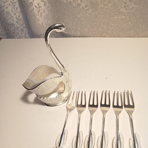 May include: A silver swan-shaped serving dish with six silver forks. The swan has a long, curved neck and a bowl-shaped body. The forks have three tines and are held by clear plastic holders.