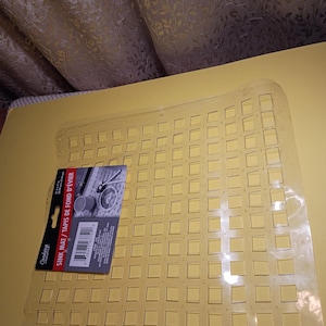 May include: A clear, flexible plastic sink mat featuring a grid of square cutouts. A product label is attached to the left side, displaying the text "Cooking Concepts," "SINK MAT / TAPIS DE FOND D'&Eacute;VIER," and dimensions of 12 in x 12 in.