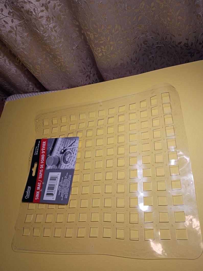 May include: A clear, flexible plastic sink mat featuring a grid of square cutouts. A product label is attached to the left side, displaying the text "Cooking Concepts," "SINK MAT / TAPIS DE FOND D'&Eacute;VIER," and dimensions of 12 in x 12 in.