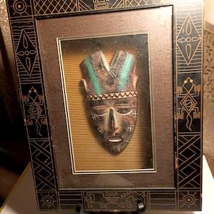 May include: Framed tribal mask with a brown and green color scheme. The mask is displayed in a dark brown frame with geometric patterns in gold. The background is a textured, golden-yellow color.