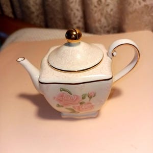 May include: A small, white porcelain teapot with a square body and a curved handle. The lid is topped with a gold knob. Pink roses decorate the front. Gold trim accents the edges. The teapot is a charming piece for tea parties or display.