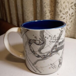 Peut inclure: Mug en c&eacute;ramique blanche avec int&eacute;rieur et anse bleus. L'illustration noire et dor&eacute;e d&eacute;taill&eacute;e de la constellation du Scorpion est imprim&eacute;e sur un fond en forme de carte. Le mug est con&ccedil;u pour les boissons chaudes.