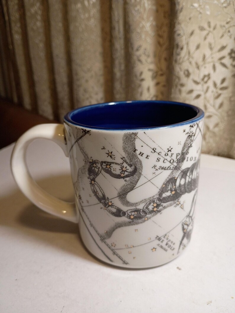 Peut inclure: Mug en c&eacute;ramique blanche avec int&eacute;rieur et anse bleus. L'illustration noire et dor&eacute;e d&eacute;taill&eacute;e de la constellation du Scorpion est imprim&eacute;e sur un fond en forme de carte. Le mug est con&ccedil;u pour les boissons chaudes.