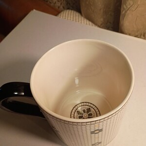 May include: A white ceramic mug with a black handle. The mug features a geometric pattern in gray and black, with a decorative design at the bottom. The interior is white, and the handle is glossy black.