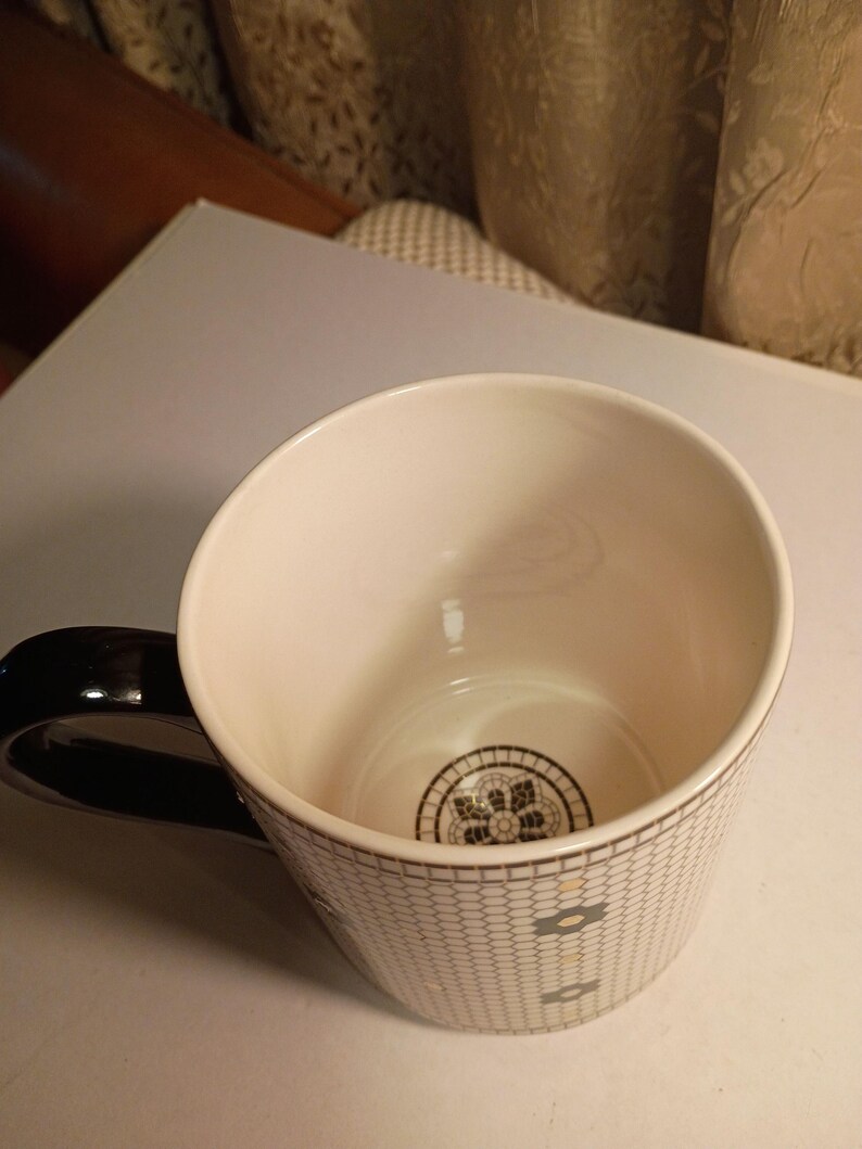 May include: A white ceramic mug with a black handle. The mug features a geometric pattern in gray and black, with a decorative design at the bottom. The interior is white, and the handle is glossy black.